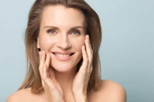Close-up portrait of smiling woman with smooth skin and bare shoulders, touching face gently, representing results of RF microneedling for skin rejuvenation